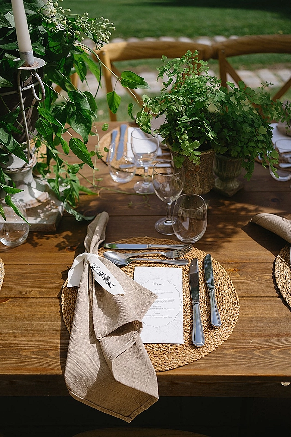 Reception tablescape with rustic wedding table setting on a wood farm table, rattan placemats, ferns, taper candles, and cross-back chairs on a lawn