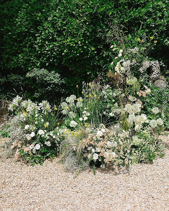 Wedding ceremony florals with ceremony aisle flowers in white, blush, and yellow wildflowers lining a gravel aisle by a garden hedge