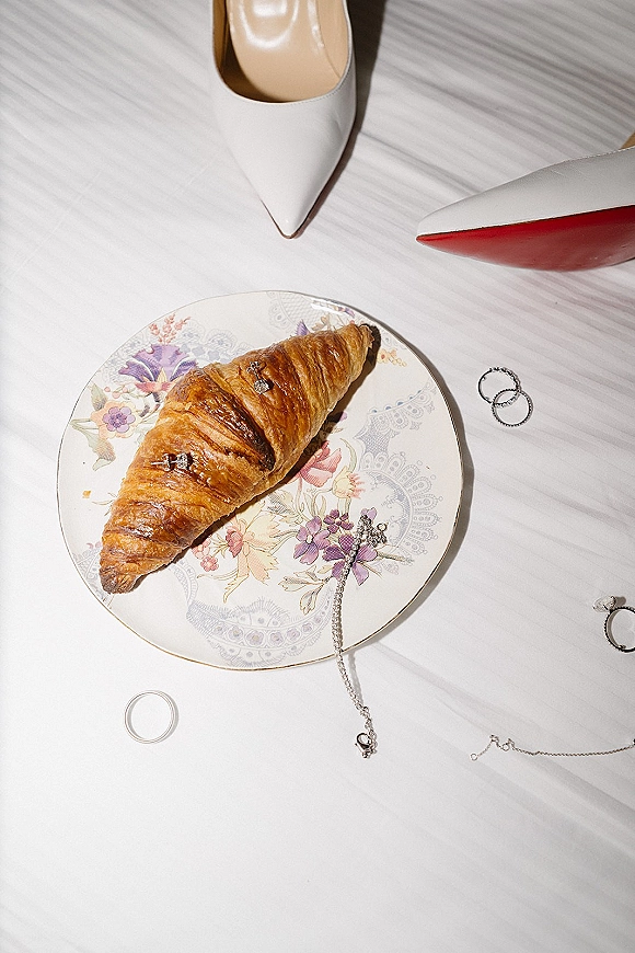 Wedding details flatlay with bridal accessories flatlay, white pointed-toe heels, rings and jewelry beside a croissant on floral plate in sun shadows