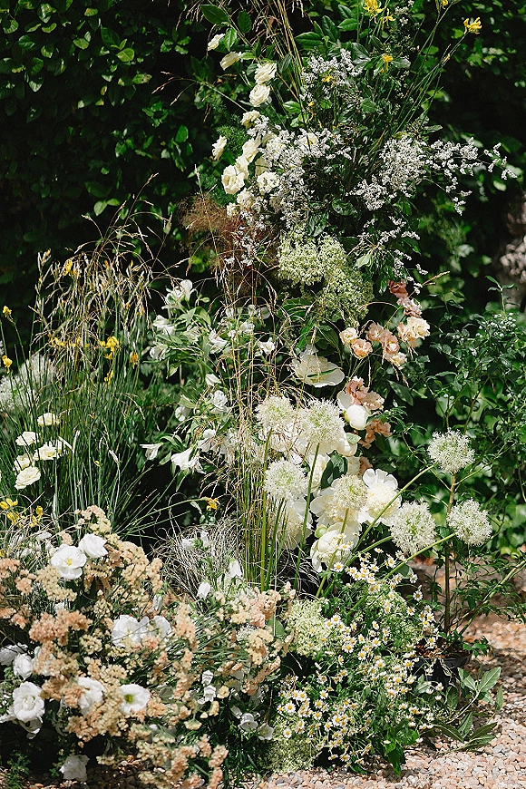 Wedding ceremony florals in a grounded ceremony floral installation with white roses, baby's breath, wildflowers and greenery on gravel in a garden setting