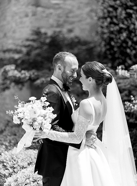 Couple portrait in a black and white wedding portrait as bride and groom embrace in profile, her veil and bouquet against garden hedges