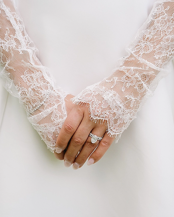 Engagement ring with an oval engagement ring on a hand, pavé band framed by lace long sleeves and neutral manicure on white fabric backdrop