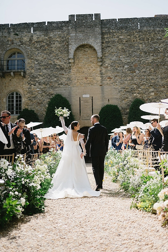 Wedding recessional as bride and groom walk away, bride raising bouquet overhead, with guests under white parasols in a castle courtyard