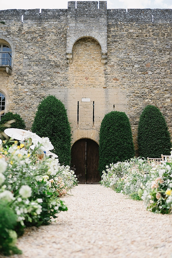 Ceremony aisle decor with garden ceremony aisle flowers lining a gravel path, parasols over wooden chairs by a stone wall and arched doorway.