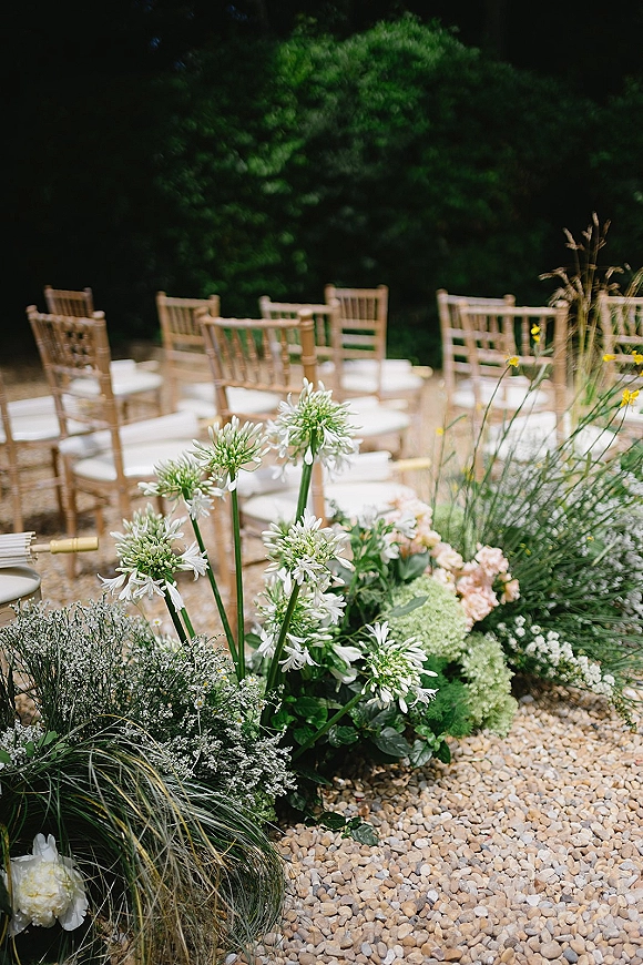 Ceremony aisle flowers with white florals and greenery in a ground arrangement beside wooden chairs with white cushions on a gravel path