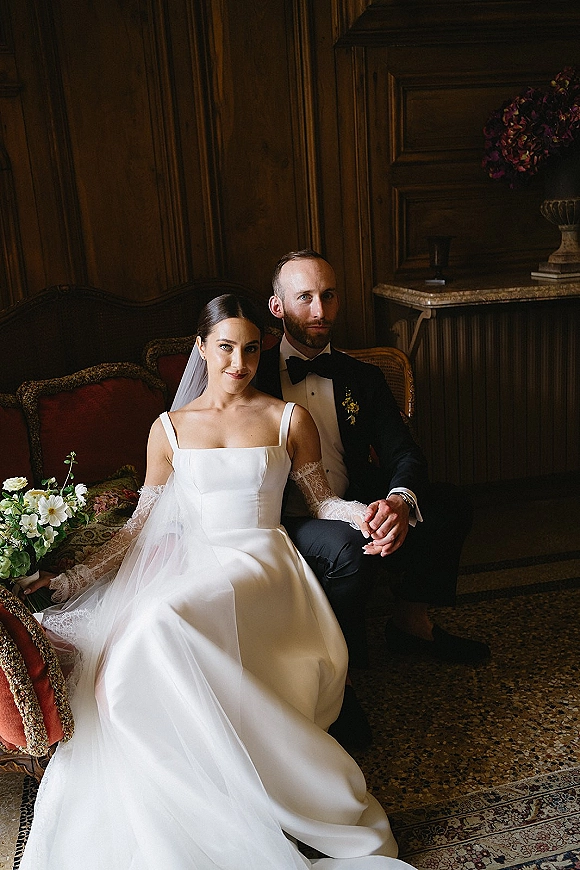 Couple portrait of bride and groom seated on a vintage sofa, holding hands, with cathedral veil and bouquet against wood paneling