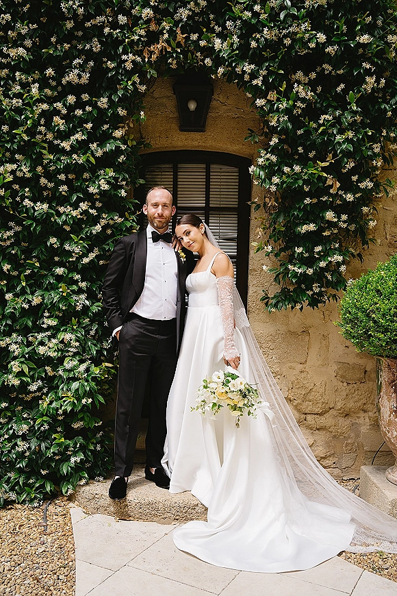Couple portrait of bride in lace sleeves and cathedral veil holding a bouquet beside groom in tuxedo by a jasmine stone wall window