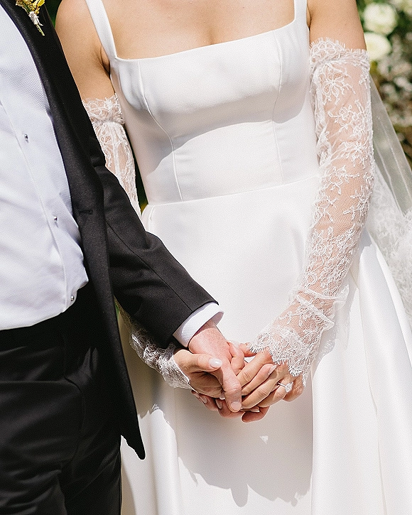 Ceremony moment of bride and groom holding hands, wedding dress lace gloves and rings in focus against greenery and white florals