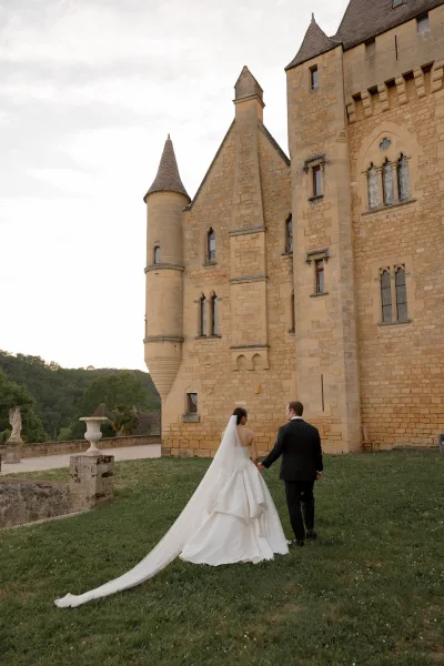 A bride and groom walk hand in hand, her cathedral veil flowing behind them against a stone castle exterior