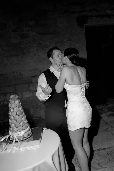 Newlyweds kiss beside a croquembouche cake on a cloth-covered table, the bride wearing a short lace dress