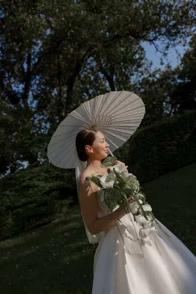 Bride holds a paper parasol beside her cascading orchid bouquet, framed by clipped garden hedges