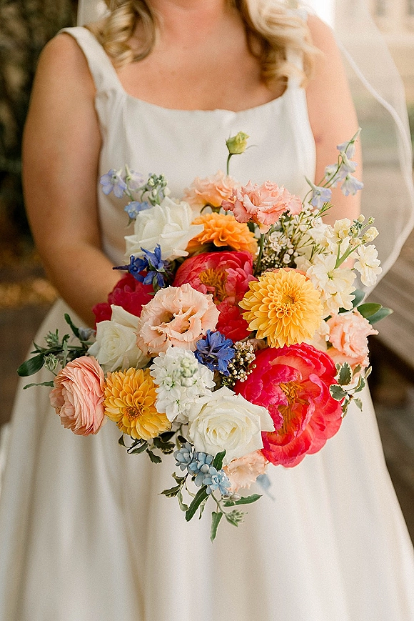Bridal bouquet, colorful wedding bouquet held against a white wedding dress with veil detail, framed by outdoor greenery in soft light