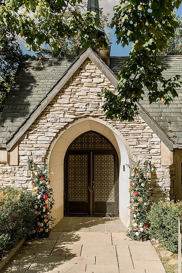 Ceremony entrance decor with chapel entrance flowers framing arched double doors, lush greenery garlands and colorful blooms on a stone chapel facade