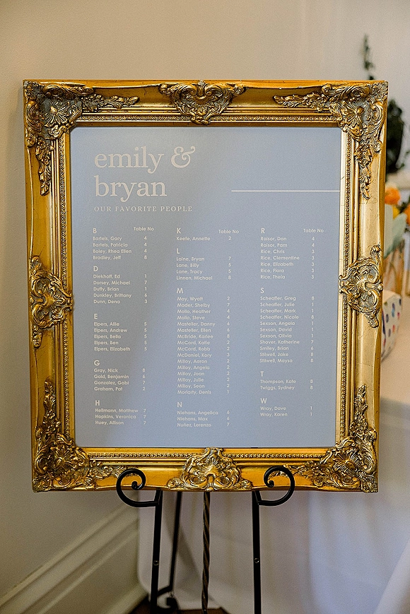 Wedding seating chart in an ornate gold frame displayed on a black metal easel against a white wall with blurred decor behind