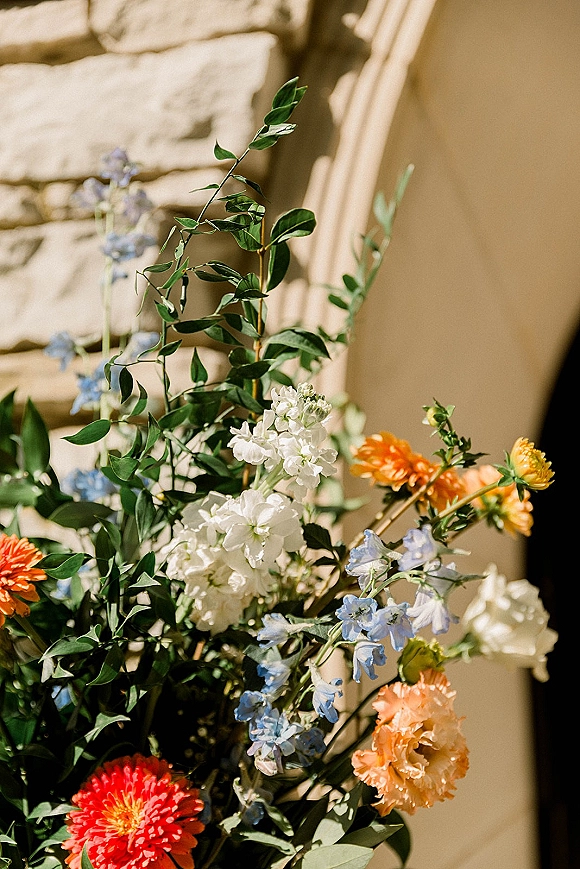 Wedding floral arrangement with colorful wedding flowers and greenery, mixing orange, blue, red, and white blooms against a sunlit stone wall