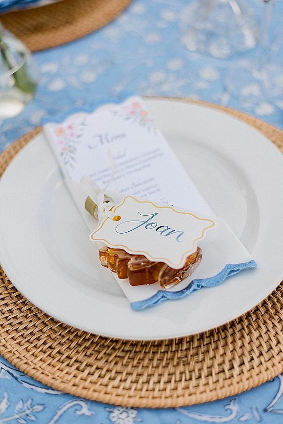 Wedding place setting with a white plate on a rattan charger, blue toile tablecloth, scalloped napkin, menu card, and caramel favor on tabletop