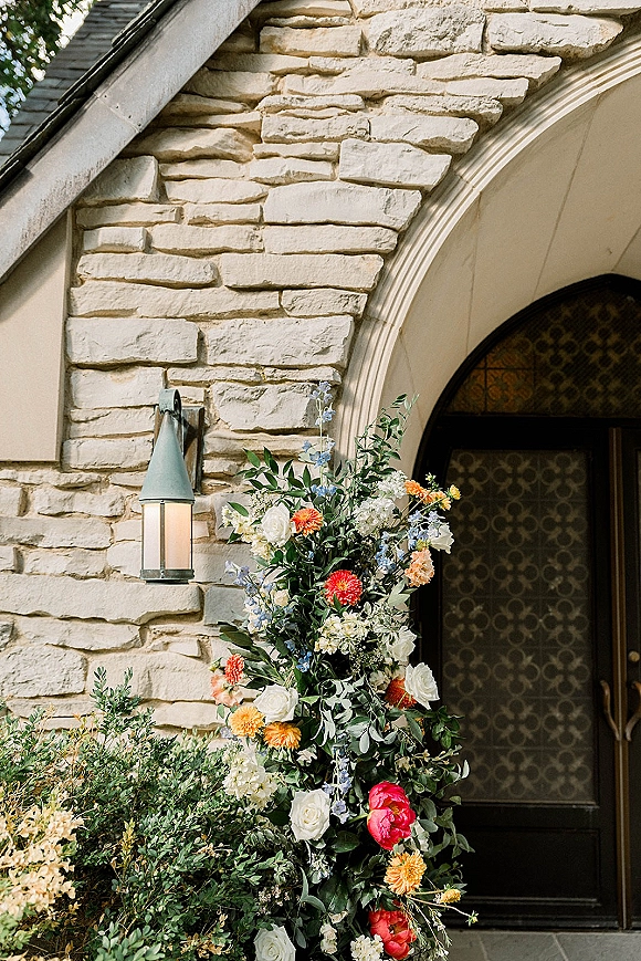 Floral entrance decor with wedding entrance flowers of roses, peonies, orange blooms and blue delphinium beside a wall lantern on a stone archway