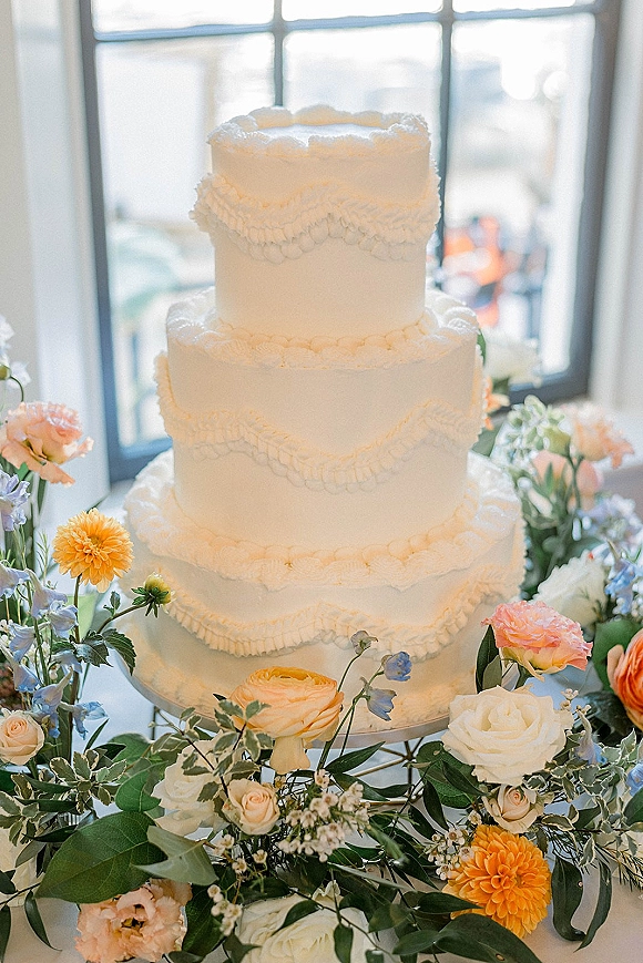 Wedding cake with three tier wedding cake design, piped ruffle buttercream on a stand with colorful florals by bright window panes