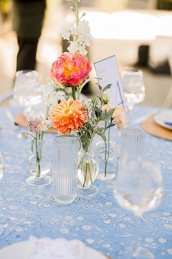 Reception tablescape with wedding bud vase centerpiece of colorful flowers in glass vases, ribbed tumblers and place card on blue toile cloth outdoors
