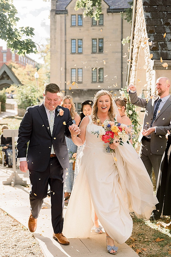 Wedding recessional as bride and groom walk hand in hand through a confetti toss exit outside a stone church, bouquet and veil flowing behind them