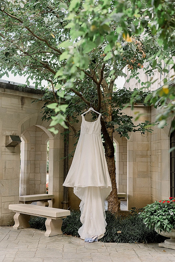 Wedding dress on hanger beneath a tree, with bridal shoes below, set in a stone courtyard with arched windows and greenery
