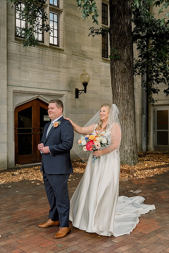 Wedding first look as bride tapping groom shoulder, veil and colorful bouquet flowing behind her in a brick courtyard by stone doors