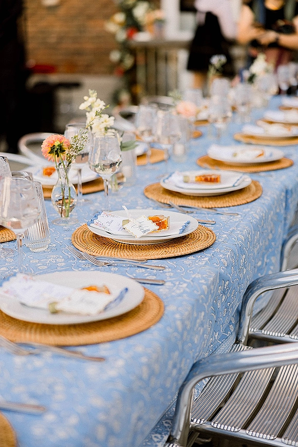 Reception tablescape with blue tablecloth wedding details, rattan placemats, gold-rim plates, bud vases, and place cards on a brick patio
