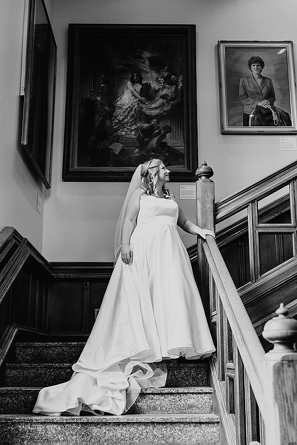 Bridal portrait of a bride in a strapless wedding dress and long veil on a staircase, framed paintings behind her in a stairwell