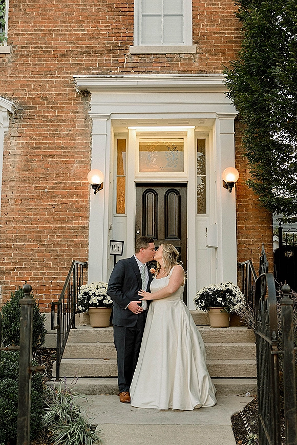 Wedding kiss portrait of bride and groom kissing on stone steps, her satin gown against his navy suit by a brick front door entrance