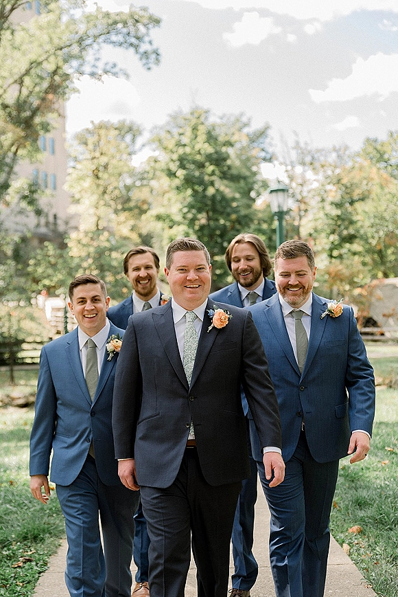 Groomsmen portrait of the groom with groomsmen walking in navy suits with boutonnieres along a park path lined with trees