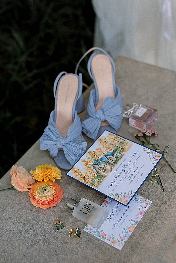 Invitation suite wedding invitation flat lay with RSVP card, rings, bridal shoes, perfume, and flowers on a stone surface with greenery