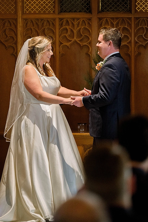 Ceremony moment as bride and groom holding hands during wedding vows, her cathedral veil flowing in an indoor space with wood panels