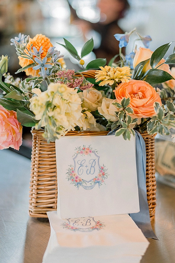 Wedding invitation suite with floral monogram invitation, wax seal envelope and ribbon beside greenery and wicker basket on a table