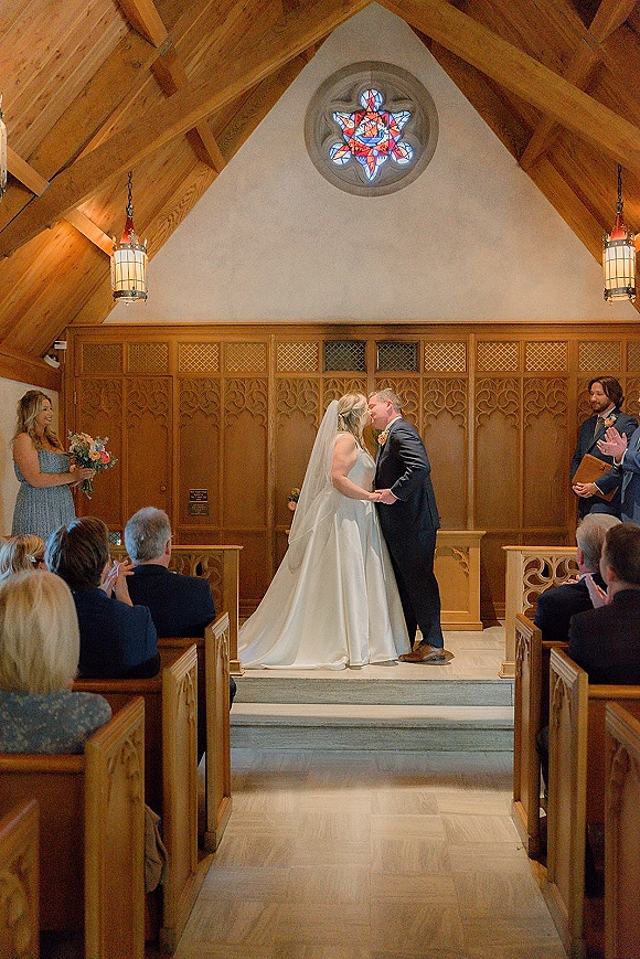 Ceremony kiss as bride in veil and satin dress kisses groom in navy suit at chapel altar, stained glass window behind, guests in pews
