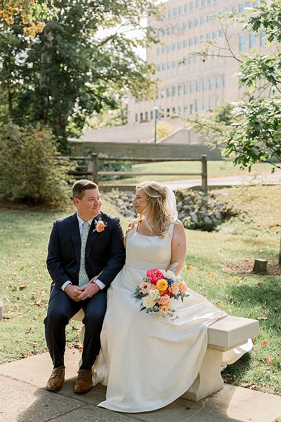 Couple portrait of bride and groom sitting on a bench, bride holding a bouquet with veil accents on a grassy lawn near trees and a fence