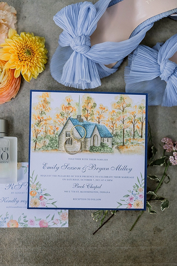 Wedding invitation suite with watercolor wedding invitation, rings, blue bow shoes and perfume arranged on gray fabric with floral sprigs
