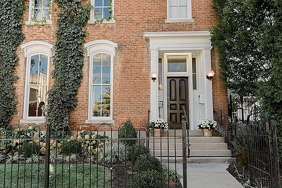 Wedding venue exterior with ivy on a brick facade, black front door and front steps framed by potted white flowers and wrought iron fence.