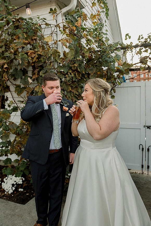 Wedding couple toast as bride in strapless satin gown and groom in navy suit clink glasses on ivy-wall patio under string lights