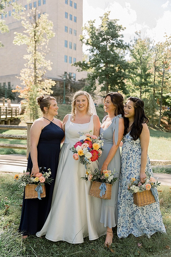 Bridesmaid group portrait with bride with bridesmaids holding wicker baskets of wildflowers tied with blue ribbons on a sunny park lawn