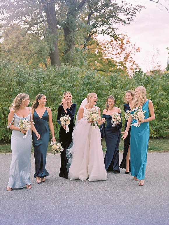 Bridesmaid group photo of bride and bridesmaids walking with white and blush bouquets, bride’s veil flowing beside a green hedge outdoors