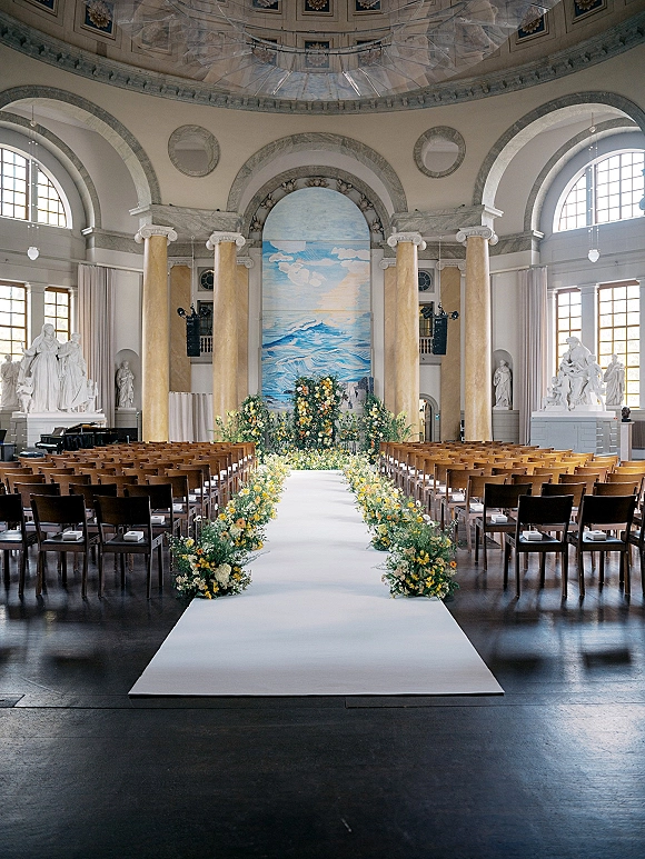 Ceremony aisle design with a white aisle runner, flanked by floral arrangements leading to an altar in a grand hall with arched windows