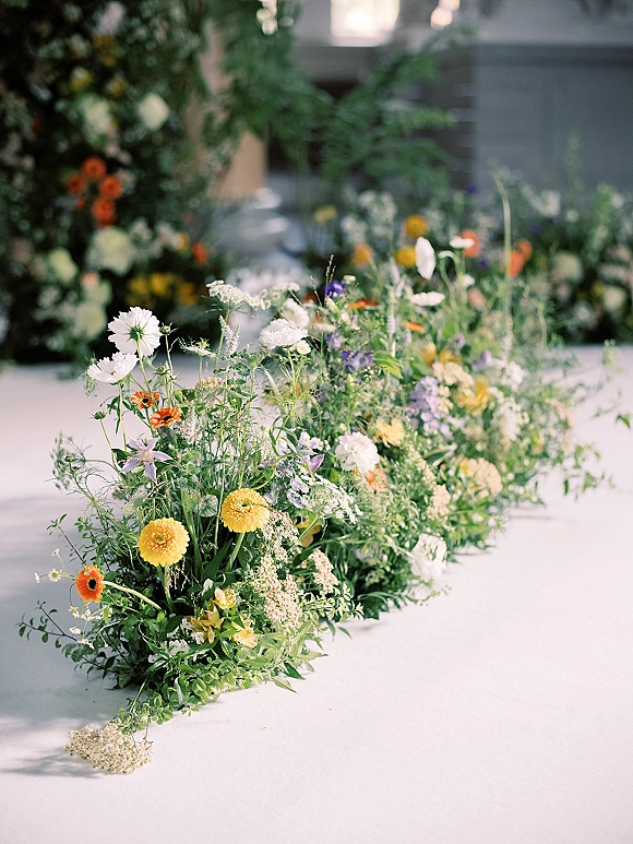 Ceremony aisle flowers create a wedding aisle floral runner of meadow wildflowers and greenery garland along a white floor with blurred greenery backdrop