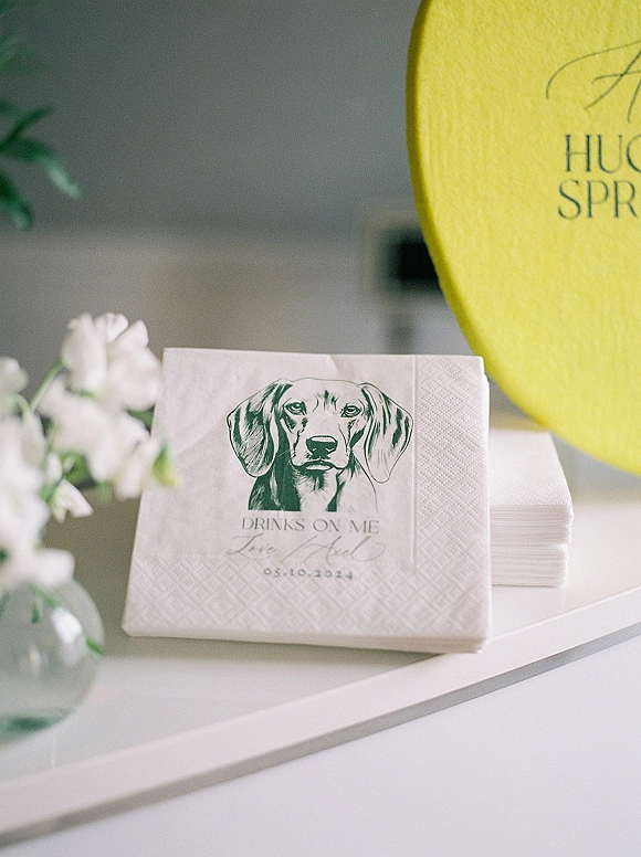 Cocktail napkins, custom cocktail napkins stacked with green dog illustration beside flowers in a glass vase on a white tabletop with balloon