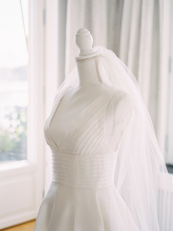 Wedding dress on a dress form mannequin with pleated bodice and satin waistband, tulle veil draped in soft window light by sheer curtains