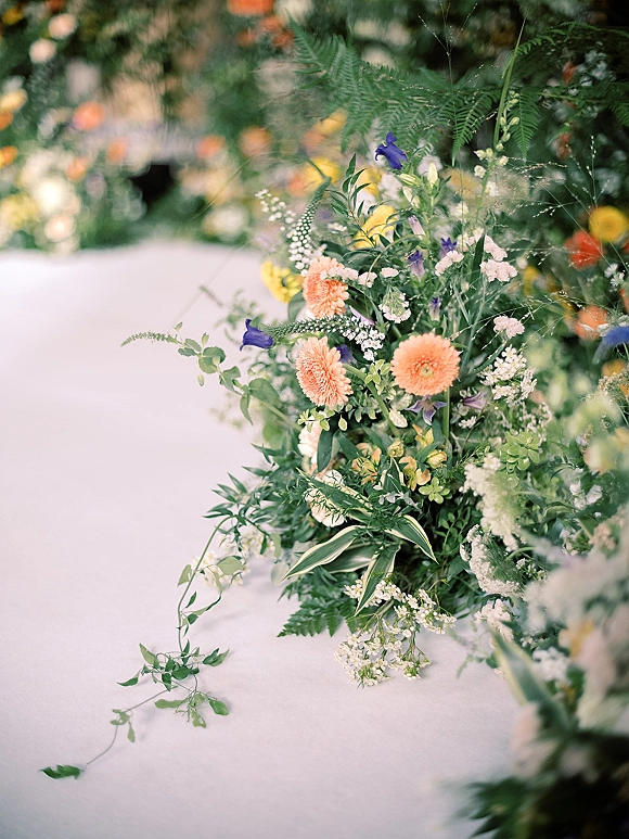 Wedding aisle florals with ceremony aisle flowers lining a white runner, featuring peach blooms, purple flowers, ferns, and baby's breath in a garden setting