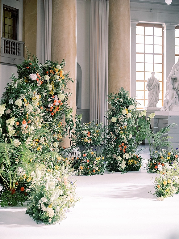 Ceremony floral decor with wedding ceremony flowers lining a white aisle runner, wildflower greenery in a grand hall with columns and statues