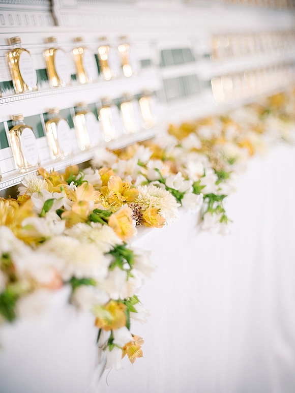 Wedding escort cards in gold place card holders lined alphabetically on a shelf with a white and yellow floral garland against a white wall