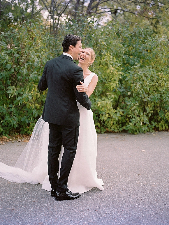 Couple portrait of bride and groom embrace, laughing as they hold hands on a tree-lined path, her veil trailing beside his black tuxedo