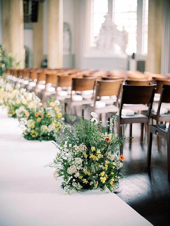 Ceremony aisle decor with a wedding aisle runner, wildflower arrangements and greenery beside wooden chairs in an indoor space with tall windows