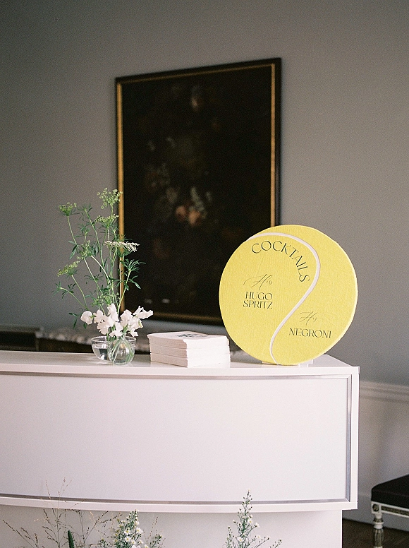 Wedding cocktail sign with signature cocktail menu on an acrylic stand beside a bud vase of white flowers on a bar counter, framed art behind